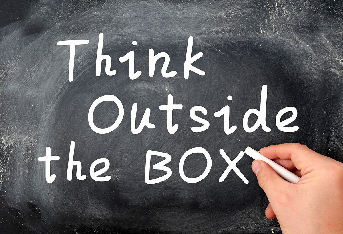 Think outside the box written on a Blackboard background with a hand holding chalk