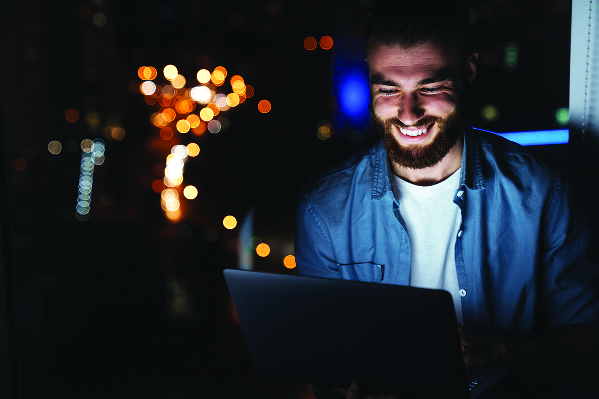 Happy young man working on laptop computer while standing in front of the window indoors at night